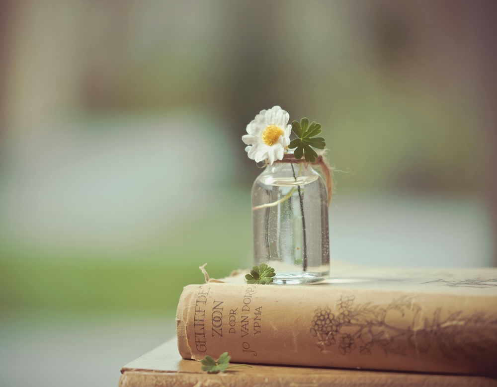 Small glass bottle with a white flower resting on top of stacked books